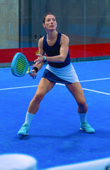 A determined female padel player prepares just before a powerful forehand, her eyes fixed on the approaching ball on a blue indoor court.