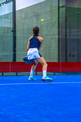 Obraz premium Rear view of a padel player watching the ball after it bounces off the glass wall preparing for an overhead shot on a blue indoor court.
