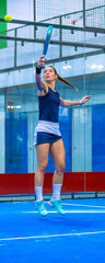 Full image of an athletic female padel player leaping for a spectacular aerial smash on a blue indoor padel court.