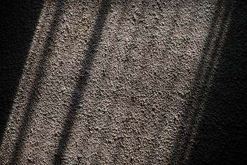 Rough stucco wall illuminated by sunlight casting diagonal grid shadows. The high-contrast light accentuates the deep granular texture of the concrete surface, creating a geometric abstract patter
