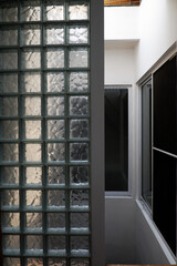 Vertical architectural view of a translucent glass block wall adjacent to a narrow residential light well. Natural daylight illuminates the white walls and aluminum window frames of the multi-story vo