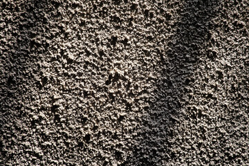 Rough stucco wall illuminated by sunlight casting diagonal grid shadows. The high-contrast light accentuates the deep granular texture of the concrete surface, creating a geometric abstract patter