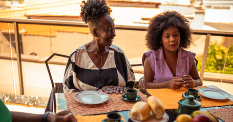Happy Black grandmother and granddaughter share a joyful conversation over a meal on a sunny balcony, enjoying a special family moment together.