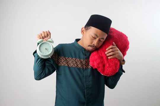 Sleepy Asian Muslim man in a green traditional outfit hugging a red pillow and holding an alarm clock, looking drowsy after suhoor during Ramadan fasting. Isolated on a white background.
