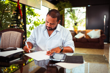 Focused man diligently writing on documents at a glass table in a bright, modern interior with a festive Christmas tree in the blurred background.