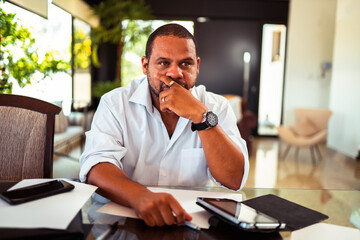 Thoughtful Black businessman sits at a modern glass desk, contemplating ideas with a smartphone and papers in a bright, contemporary office.