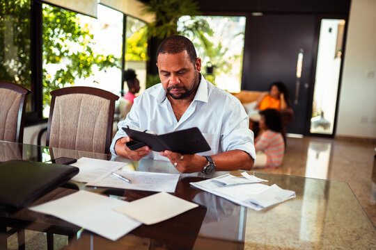 Focused African American man reviews documents and manages finances at a glass table in his modern home, with natural light and blurred family in the background. - Powered by Adobe