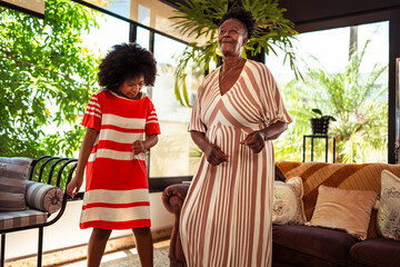 Happy African American grandmother and granddaughter dancing together in a bright, plant-filled living room.
