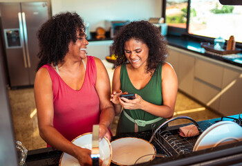Smiling Black women share a joyful moment in a modern kitchen, one washing dishes while the other uses a smartphone.