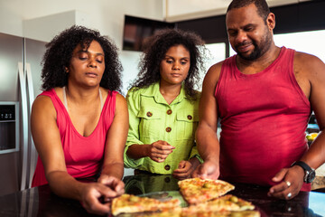 Three diverse people gather in a modern kitchen, reaching for slices of delicious pizza on the countertop.