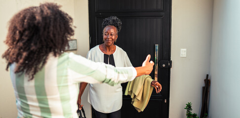 Happy African American woman welcomes an older relative with open arms at the front door of her home, showing warmth and family connection.