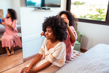 African American mother styling her daughter's natural curly hair with a comb in a bright bedroom,...