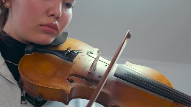 Close-up shot of a young Asian student practicing the violin at home. Focused expression, precise bow movement and clear view of the wooden instrument. Music lesson or private practice session.