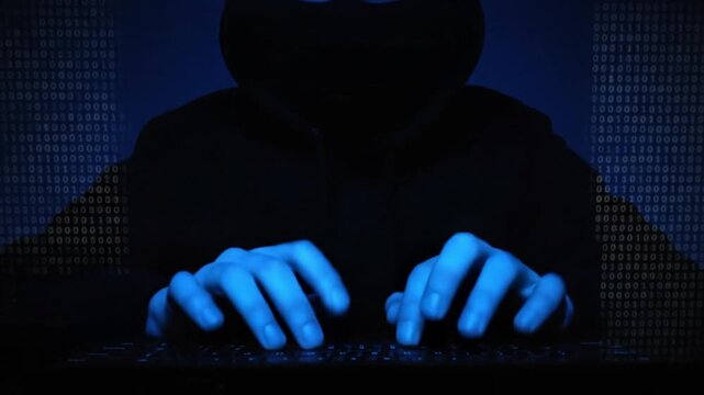 A mysterious, hooded figure is intensely focused on typing on a computer keyboard in a dimly lit environment. The scene is bathed in a dramatic blue light, highlighting the hands and keycaps, while a 