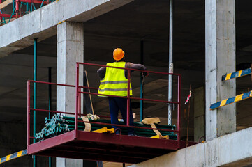 Construction worker standing on a raised platform at a building site. The worker is wearing a yellow high-visibility safety vest and an orange hard hat.