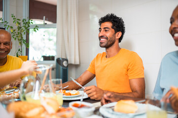 Happy young man in orange t-shirt enjoying a meal with family and friends at home. Diverse group of...
