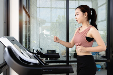 Fototapeta premium Asian happy beautiful woman running on a treadmill in sport gym. Side profile young fitness girl engaging in jogging workout exercises, training on treadmill machine