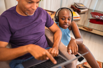 African American couple using laptop together at home, woman with headphones pointing at screen.