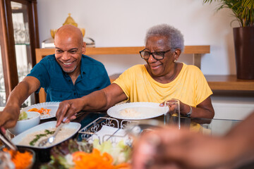 Happy elderly Black woman and adult son sharing a healthy meal together at home, smiling and serving food during a family lunch.