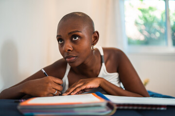 Pensive young African American woman with a shaved head writing in her journal while lying down at home, looking up thoughtfully. © Nexa
