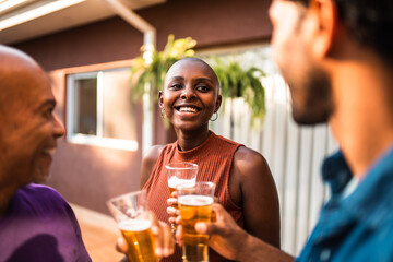 Smiling African American woman with shaved head enjoying a beer with family and friends at an outdoor backyard gathering during golden hour.