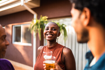 Happy Black woman with a shaved head smiling and holding a beer at an outdoor backyard party with friends.