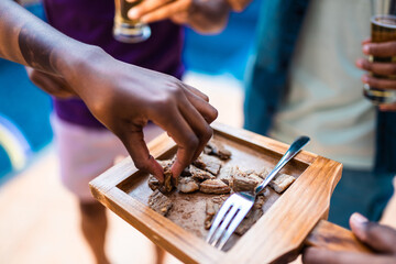 Close-up of hands taking grilled meat from a wooden board at an outdoor party with friends.