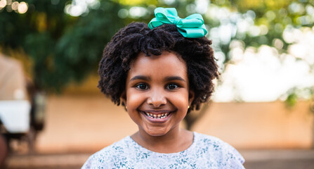 Portrait of a happy young Black girl with curly afro hair and a turquoise bow smiling outdoors.