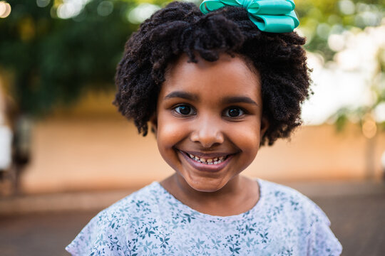Portrait of a joyful young Black girl with curly hair and a teal bow smiling at the camera outdoors.