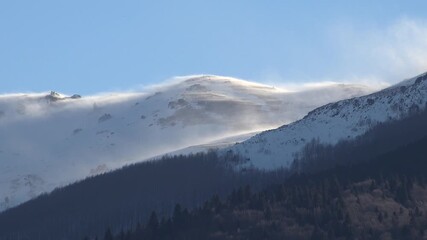 Strong blizzard wind sculpts snow surface across high mountain slope under clear winter sky. Surface erosion patterns reveal harsh alpine conditions during sunny cold weather.