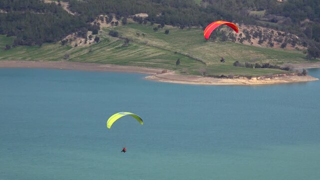 Colorful paragliders soar above calm lake near forested shoreline during outdoor adventure flight. Bright wings and open sky create dynamic leisure sport scenery.