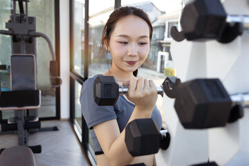 Fototapeta premium Asian smiling beautiful woman lifts dumbbell from dumbbell rack to exercise in sport gym. Young fitness girl engaging in workout exercises, training to keep body strong and fit, healthy bodybuilding.
