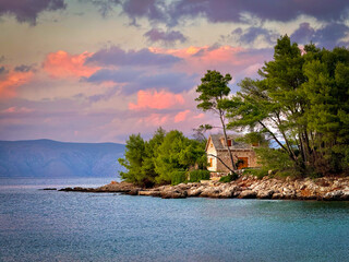 Rustic stone house stands by the calm sea beneath soft pink clouds of a colorful sunset sky. Lush pine trees and rocky shore create a tranquil Mediterranean atmosphere in hidden bay on island of Hvar.
