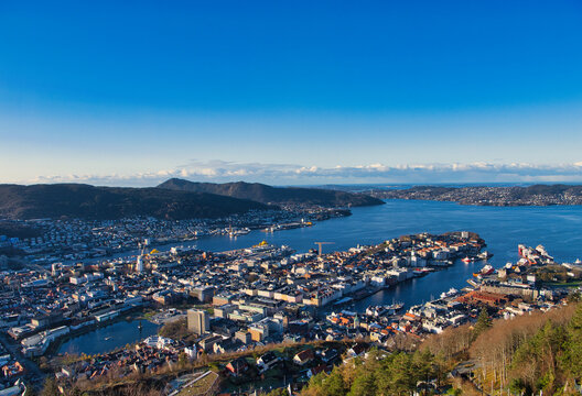 Wide panoramic view over Bergen, Norway, with the harbor, waterfront city center, and fjord under a clear blue sky. Bright, crisp daytime travel scene.