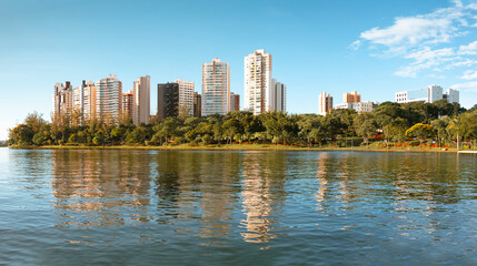 Urban landscape with modern buildings reflected in the lake, surrounded by green trees under a blue, sunny sky.