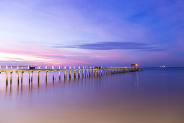 Obraz premium Long wooden pier at dusk with soft purple sky and calm water reflecting lights at Bang Pu in Samut Prakan, Thailand.