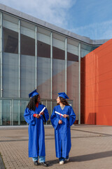 Two diverse female students wearing cap and gown smiling with diplomas on campus