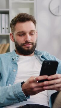 Portrait of attractive young man sitting on the sofa using phone smile in the modern apartment texting message content on smartphone social media applications online scrolling tapping technology. Slow