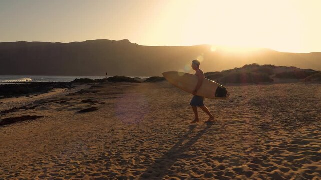 LENS FLARE: Male surfer runs to the beach with wooden surfboard tucked under his arm in golden light. He stops before heading into water to check breaking waves at iconic surf spot on Lanzarote island