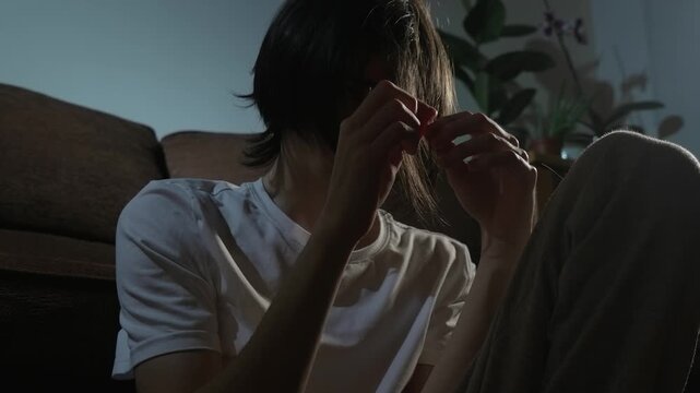 Depression weighing on young man sitting alone and nervously twisting hair in dim room. Person fidgeting with strands while sinking into heavy emotional stress. Teenager touching hair showing signs of