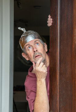 Tinfoil hat picture. A man wearing a tinfoil hat looks anxiously out of a door towards the sky. Paranoia and conspiracy theory. Alien abduction and government cover up. Mind control and brainwashing.