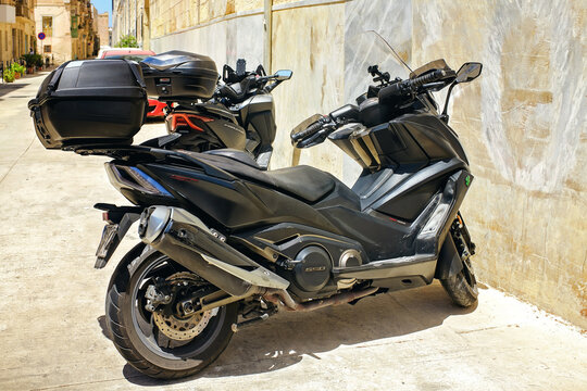 Bormla, Malta - July 13, 2025: Black SYM Maxsym TL and Honda Forza 350 scooters with top cases parked along a wall on a sunny street.