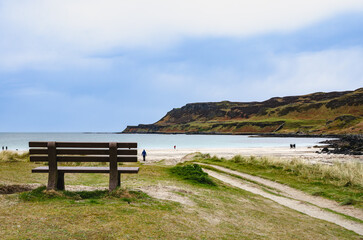 Wooden bench by a sandy coastal track on the Isle of Mull, Scotland, overlooking beach, dunes and distant cliffs. Peaceful seaside viewpoint for travel and relaxation themes.