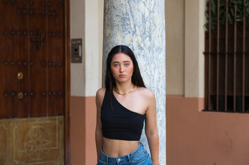A serious, young, attractive woman with dark hair, dressed in jeans and a black top, leaning back and holding onto the grey marble column in the centre of Seville with her hands.