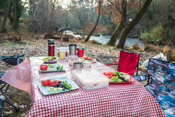 Traditional turkish breakfast picnic table with red checkered tablecloth set near a flowing river in the autumn forest