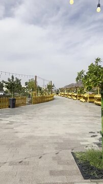 Daytime view of Suq Al-Uyun Market in Medina, near Mount Uhud, featuring yellow wooden stalls, hanging string lights, and modern stone paving, elegantly framed by a wooden fence beneath a cloudy sky.