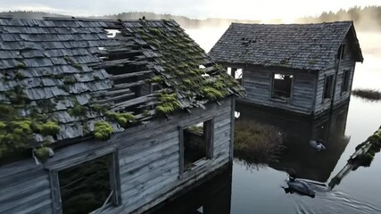Drone view of decaying wooden cabins submerged in calm lake water with morning fog