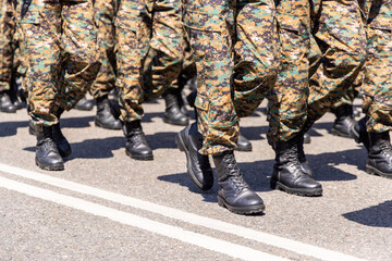A group of soldiers marches in military camouflage trousers and black boots on an asphalt road.