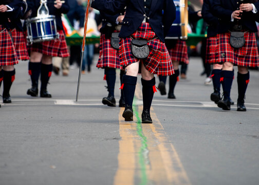 Kilt wearing bagpipe band walks down the street over a green stripe in the road during a St. Patrick's Day parade. 