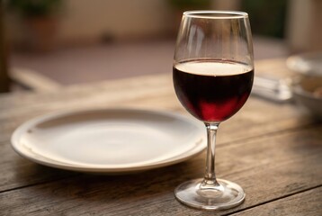 A glass filled with red wine is placed on a rustic wooden table. An empty white plate is beside the glass, suggesting a meal has taken place. It is evening time and light is soft